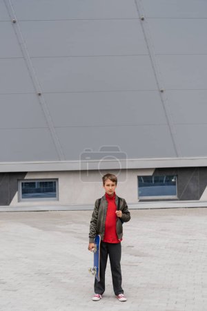 full length of preteen boy in stylish bomber jacket holding penny board near mall building 