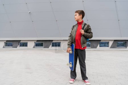 Téléchargez les photos : Full length of preteen boy in trendy bomber jacket holding penny board near mall building - en image libre de droit