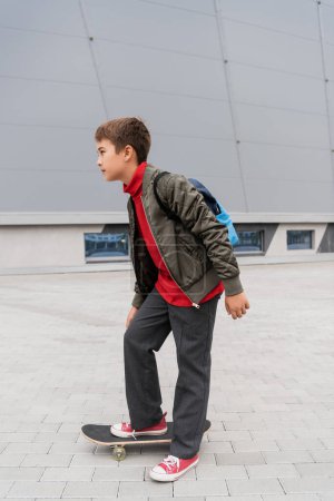 full length of preteen boy in trendy bomber jacket riding penny board near mall 