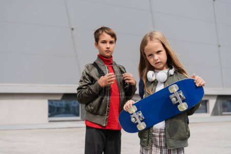 Preteen Mädchen in drahtlosen Kopfhörern hält Penny Board, während sie mit Junge in der Nähe von Gebäude von Einkaufszentrum steht 