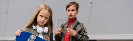 Preteen Mädchen in drahtlosen Kopfhörern hält Penny Board, während sie mit Jungen in der Nähe von Einkaufszentrum steht, Banner 