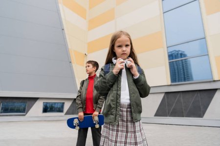 preteen girl in wireless headphones standing with stylish boy holding penny board on blurred background 