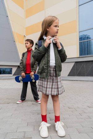 full length of preteen girl in wireless headphones standing with stylish boy holding penny board on blurred background 