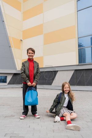 glückliches Mädchen im Rock sitzt auf Penny Board neben stilvollem Jungen mit Rucksack in der Nähe von Einkaufszentrum 