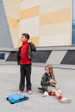 Preteen Mädchen im Rock sitzt auf Penny Board neben stilvollem Jungen in Bomberjacke in der Nähe von Einkaufszentrum 