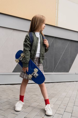 full length of stylish preteen girl in bomber jacket holding penny board standing near mall 