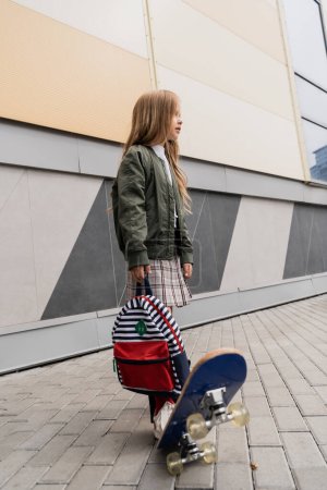 Téléchargez les photos : Full length of stylish preteen girl in bomber jacket holding backpack while riding penny board near mall - en image libre de droit