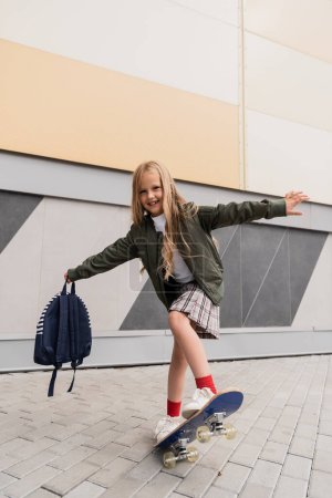Photo for Full length of happy preteen girl in stylish bomber jacket holding backpack while riding penny board near mall - Royalty Free Image