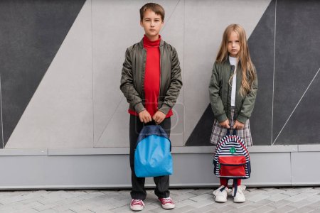 full length of stylish kids in bomber jackets holding backpacks while standing near mall 