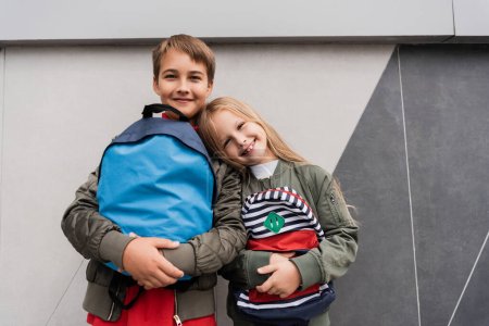Photo pour Low angle view of happy kids in trendy bomber jackets holding backpacks while standing near mall - image libre de droit