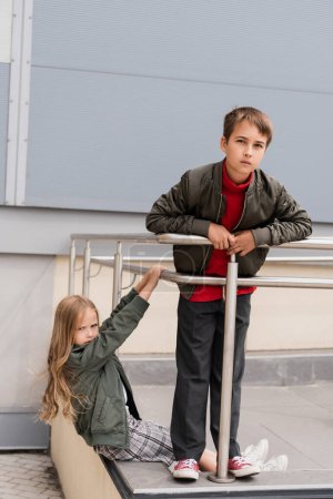well dressed preteen kids in bomber jackets posing near metallic handrails near mall 