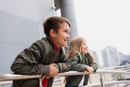happy preteen kids in bomber jackets leaning on metallic handrails near mall 