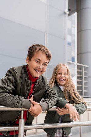 cheerful preteen kids in bomber jackets leaning on metallic handrails near mall 