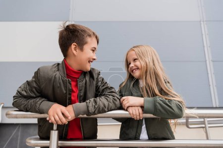 happy preteen kids in bomber jackets looking at each other while leaning on metallic handrails near mall 