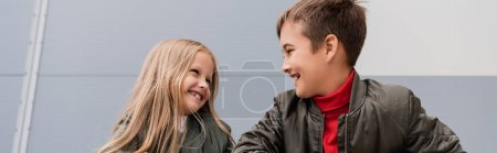 happy preteen kids in bomber jackets looking at each other while standing near mall, banner 
