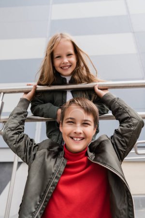 Photo for Low angle view of happy and well dressed preteen kids in bomber jackets leaning on metallic handrails near mall - Royalty Free Image