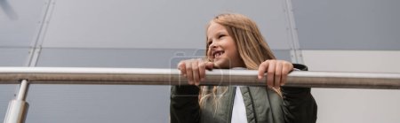 Foto de Low angle view of cheerful and well dressed preteen girl in bomber jacket leaning on metallic handrails near mall, banner - Imagen libre de derechos
