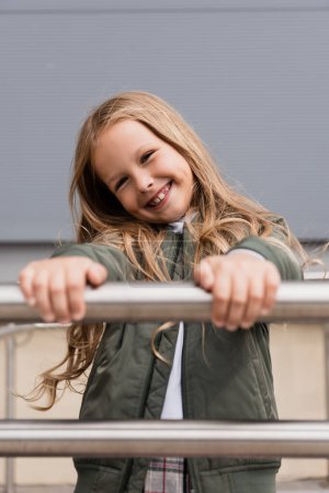 cheerful preteen girl in stylish bomber jacket leaning on metallic handrails near mall 