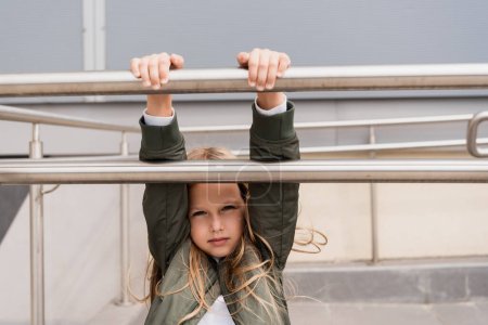 preteen girl in stylish bomber jacket leaning on metallic handrails near mall 