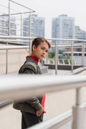 Photo for Preteen boy in stylish bomber jacket looking at camera near metallic handrails on blurred foreground - Royalty Free Image
