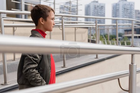 well dressed preteen boy in stylish bomber jacket standing near metallic handrails on blurred foreground 