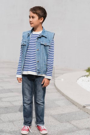 full length of well dressed boy in striped long sleeve shirt and denim vest standing outdoors 