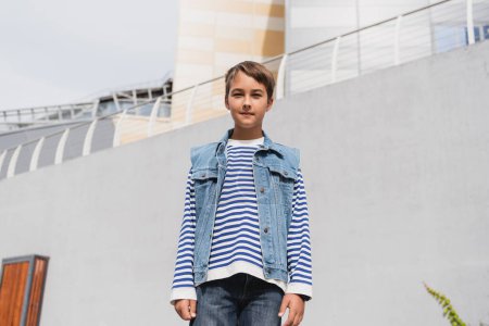 low angle view of well dressed boy in striped long sleeve shirt and denim vest standing outdoors 