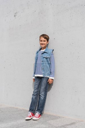 full length of happy boy in striped long sleeve shirt and denim vest leaning on wall outdoors 