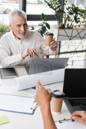 Positive middle aged businessman holding coffee to go near laptops and african american colleague in office 