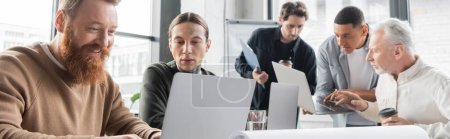 Positive businessman using laptop near tattooed colleague in office, banner 