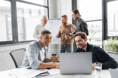 Positive multiethnische Geschäftsleute nutzen Laptop in der Nähe von Kollegen während eines Treffens im Büro 