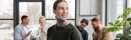 Smiling tattooed businessman standing near blurred interracial colleagues in office, banner 