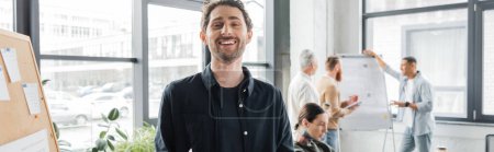 Foto de Un hombre de negocios sonriente mirando la cámara cerca del tablero y colegas borrosos en la oficina, pancarta - Imagen libre de derechos
