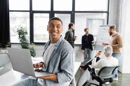Cheerful african american businessman using laptop near blurred colleagues working in office 