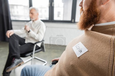 Bearded man with name sticker sitting at group therapy session in rehab center 