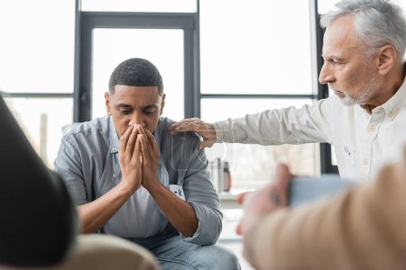Middle aged man calming african american member of alcoholics meeting in rehab center 