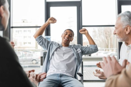 Téléchargez les photos : Homme afro-américain excité avec dépendance à l'alcool montrant un geste oui lors d'une séance de thérapie de groupe - en image libre de droit
