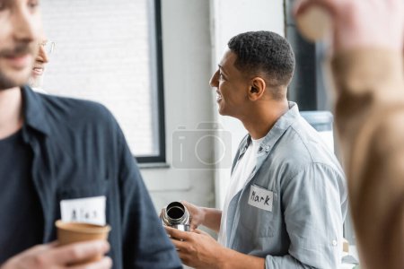 Sonriente hombre afroamericano vertiendo bebida del termo y hablando durante la reunión de alcohólicos en el centro de rehabilitación 