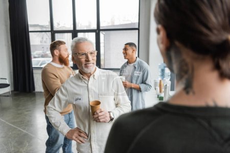 Smiling mature man with paper cup and name sticker spending time during alcoholics meeting in rehab center 