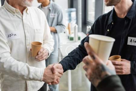 Cropped view of people holding paper cups and shaking hands during alcoholics meeting in rehab center