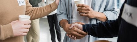 Vista recortada de personas multiétnicas con adicción al alcohol sosteniendo vasos de papel y estrechando las manos en el centro de rehabilitación, pancarta 