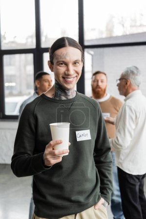 Smiling tattooed man holding paper cup and looking at camera during alcoholics meeting in recovery center 