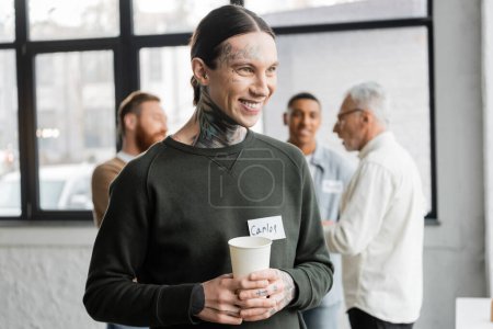 Positive tattooed man holding paper cup during meeting of alcoholics in recovery center 