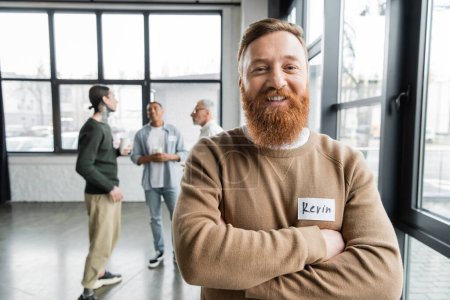 Cheerful bearded man crossing arms and looking at camera during alcoholics meeting in rehab center 