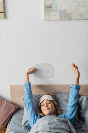 Photo for Tattooed woman in sleeping mask and nightwear stretching while lying in bed - Royalty Free Image