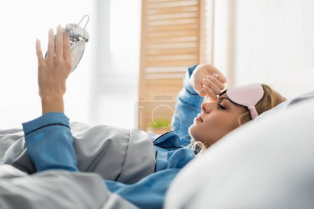 Photo for Side view of young woman in sleeping mask looking at alarm clock while lying on bed - Royalty Free Image