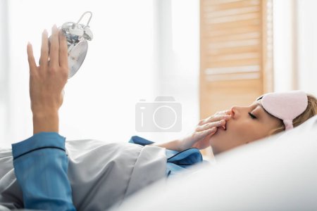 Foto de Vista lateral de la mujer joven en la máscara de dormir bostezando mientras sostiene el reloj despertador en la mañana - Imagen libre de derechos