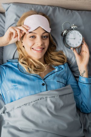 Photo for Top view of smiling woman in sleeping mask and blue pajama holding alarm clock while lying in bed - Royalty Free Image