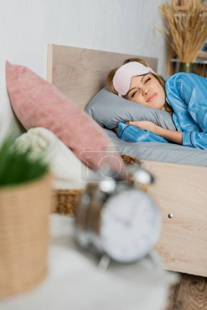 Unzufriedene Frau in Schlafmaske und Pyjama liegt neben verschwommenem Wecker im Bett 