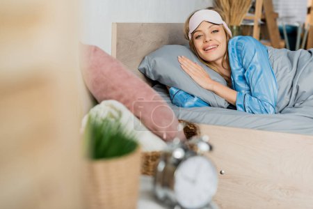 fröhliche Frau in Schlafmaske und Pyjama, die neben verschwommenem Wecker im Bett liegt 
